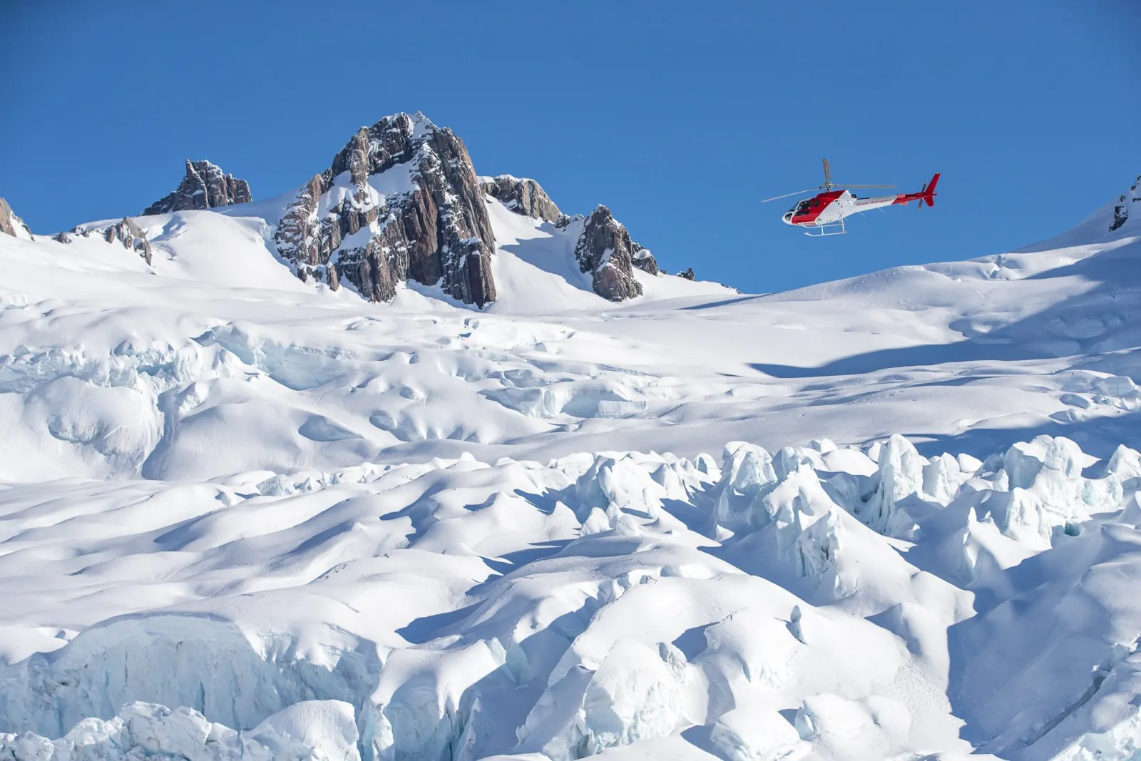 Small red helicopter flying above a crevassed snow glacier toward a rocky pinnacle