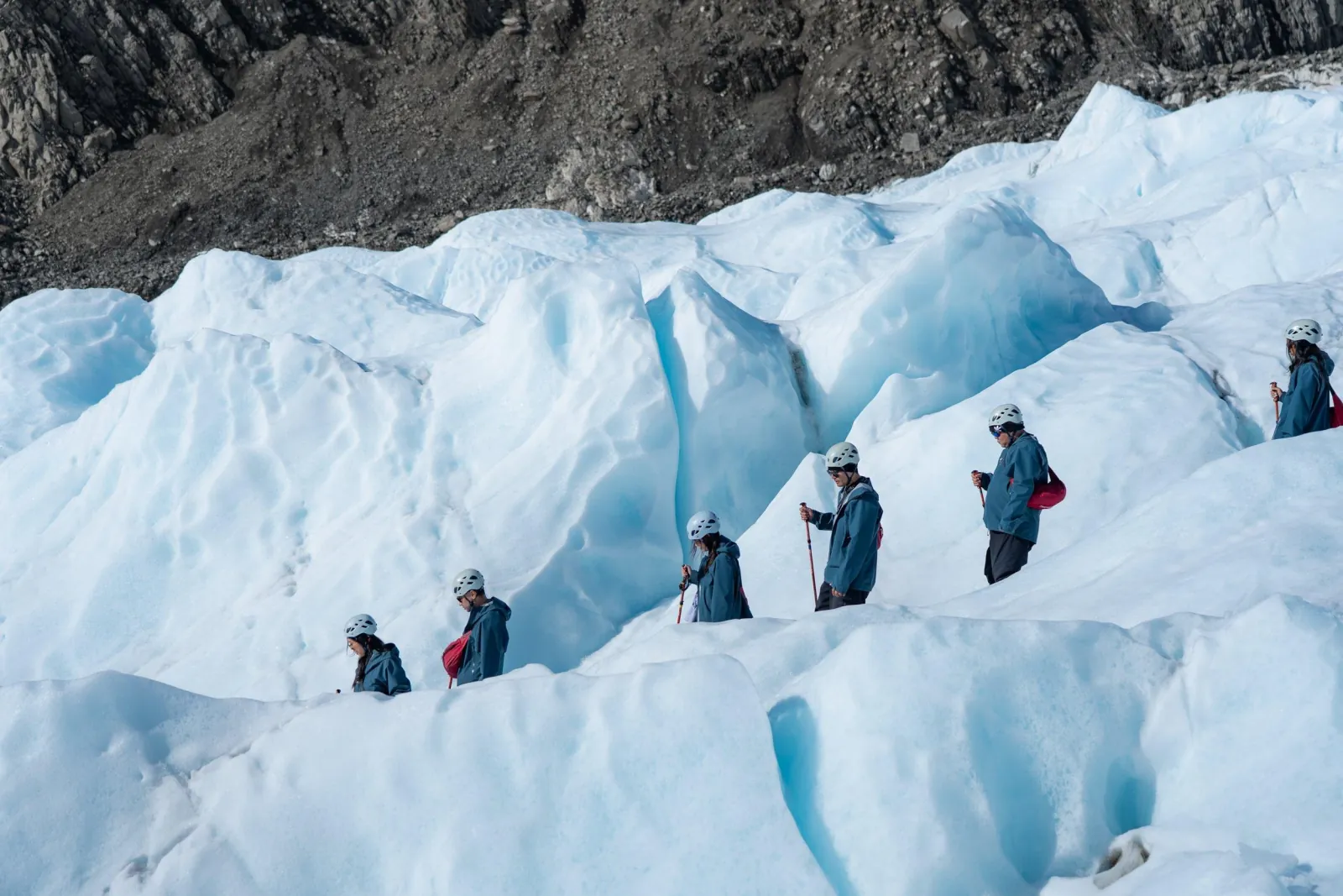 Hikers in blue jackets walking in single file along a ridge of glacier ice
