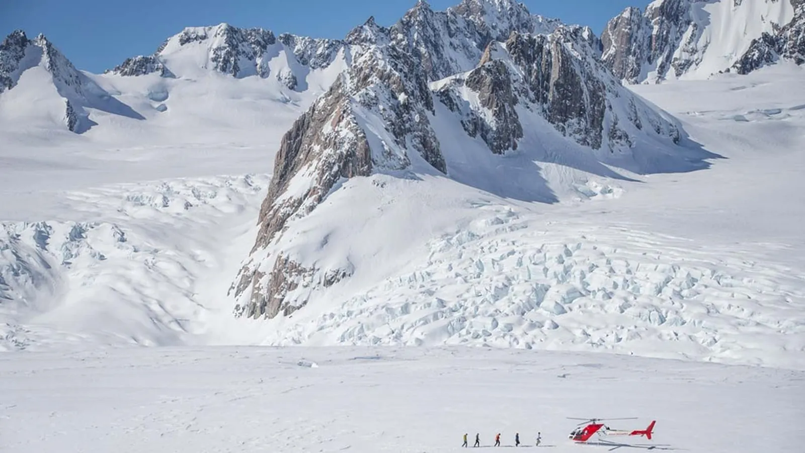 Wide snowfield below a rocky ridge with a tiny helicopter and people at the bottom