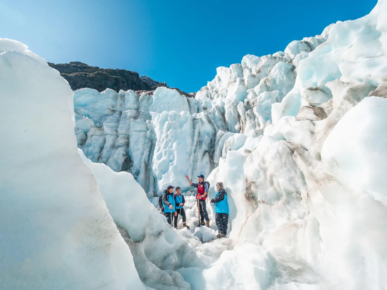 Small group of hikers standing at the foot of a tall ice wall under blue sky