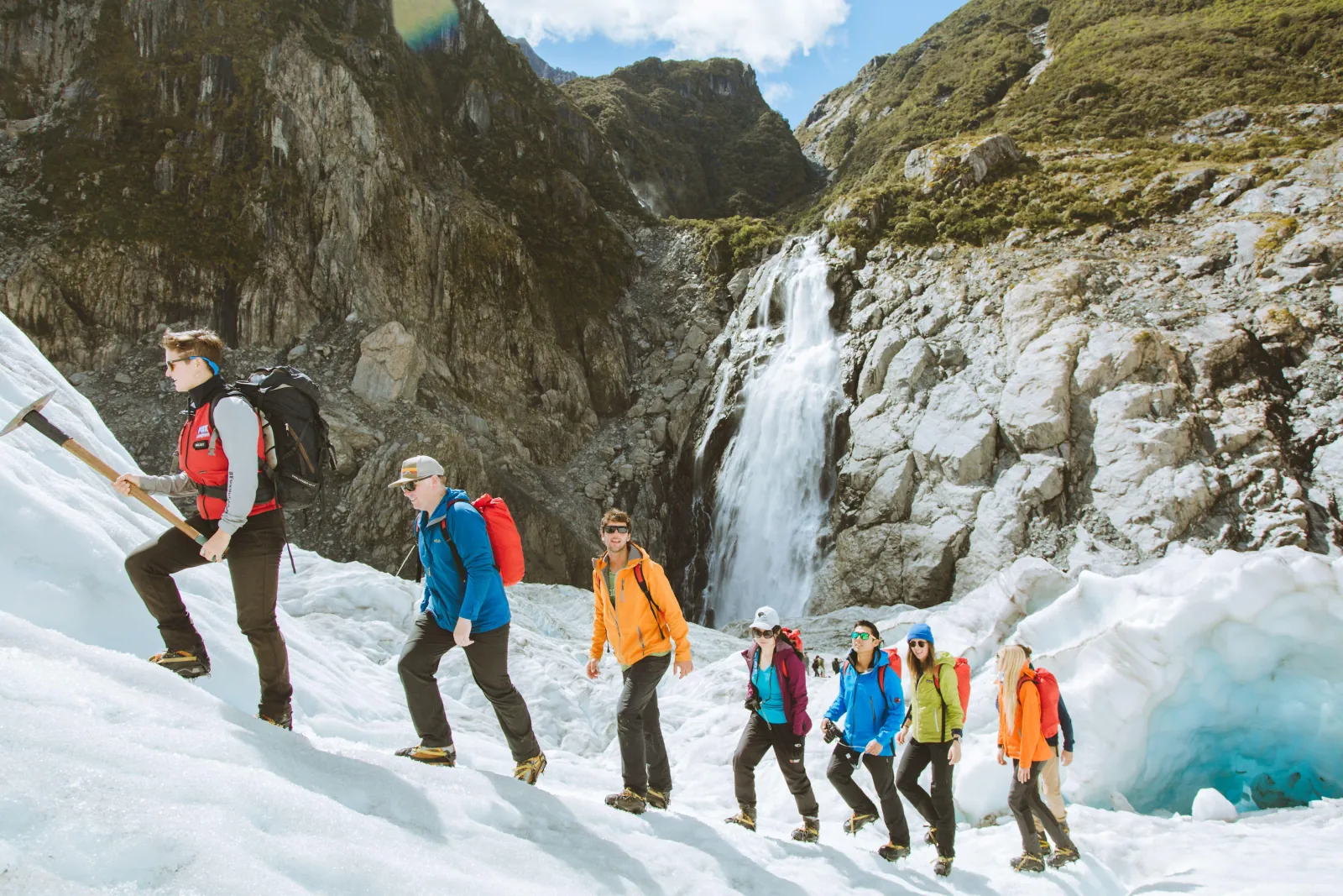 Guide and hikers in colourful jackets climbing an icy slope below a waterfall