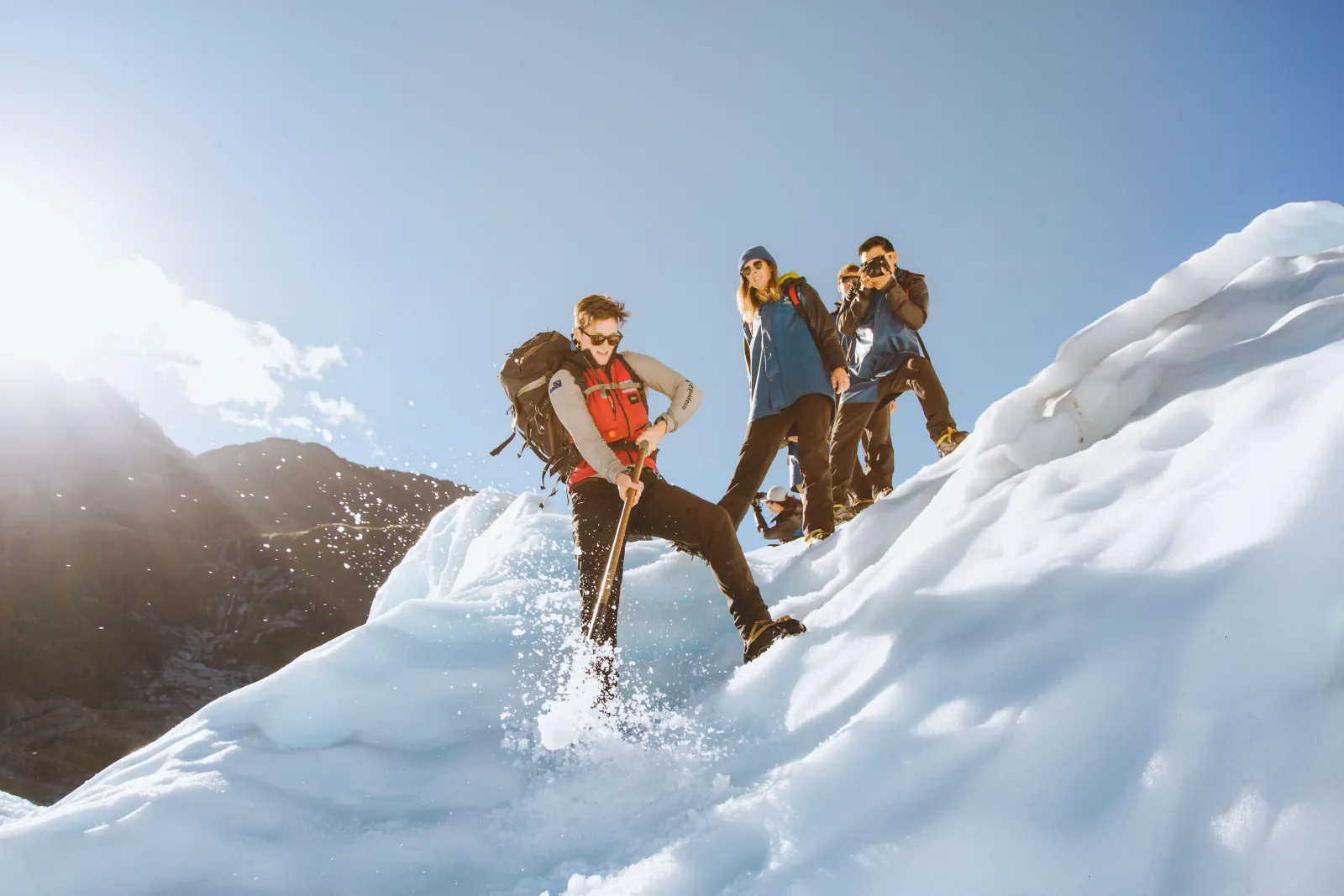 Three hikers with packs and ice axes standing on a sunlit snow ridge
