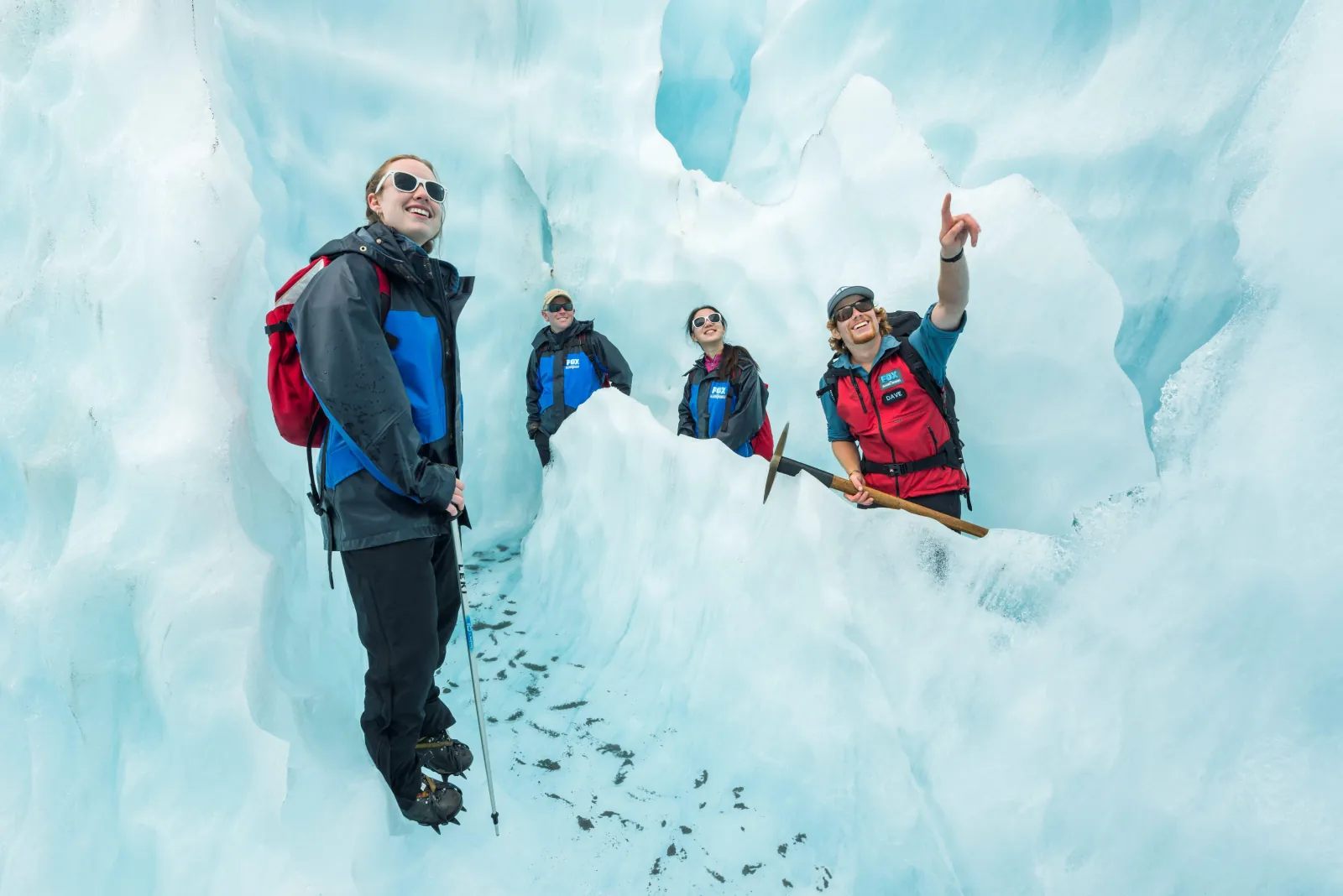 Guide pointing upward as hikers in helmets stand in a pale blue ice cave