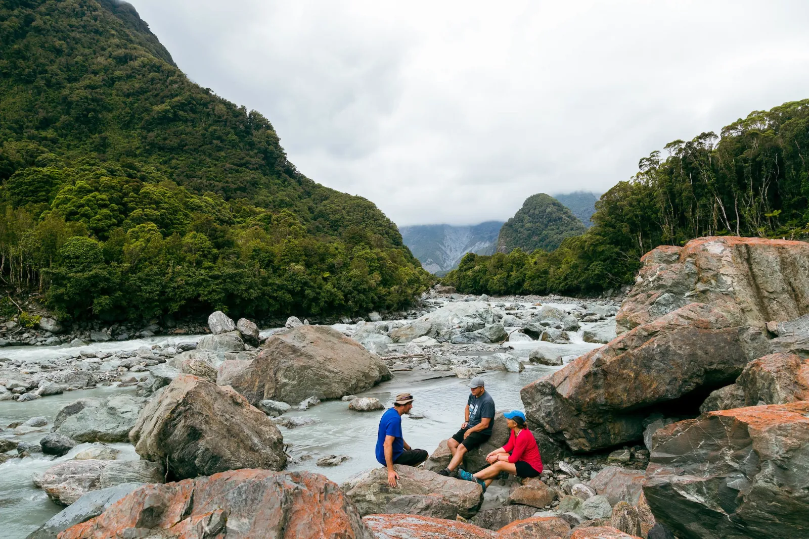 Walkers sitting on boulders beside a braided glacial river in a forested valley