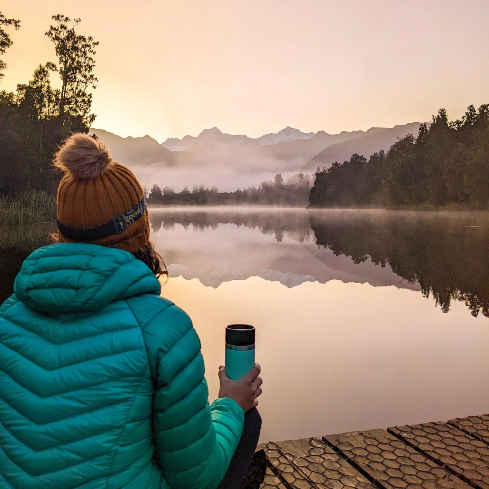 Walker in a woolly hat holding a coffee cup on a jetty over the misty lake at dawn