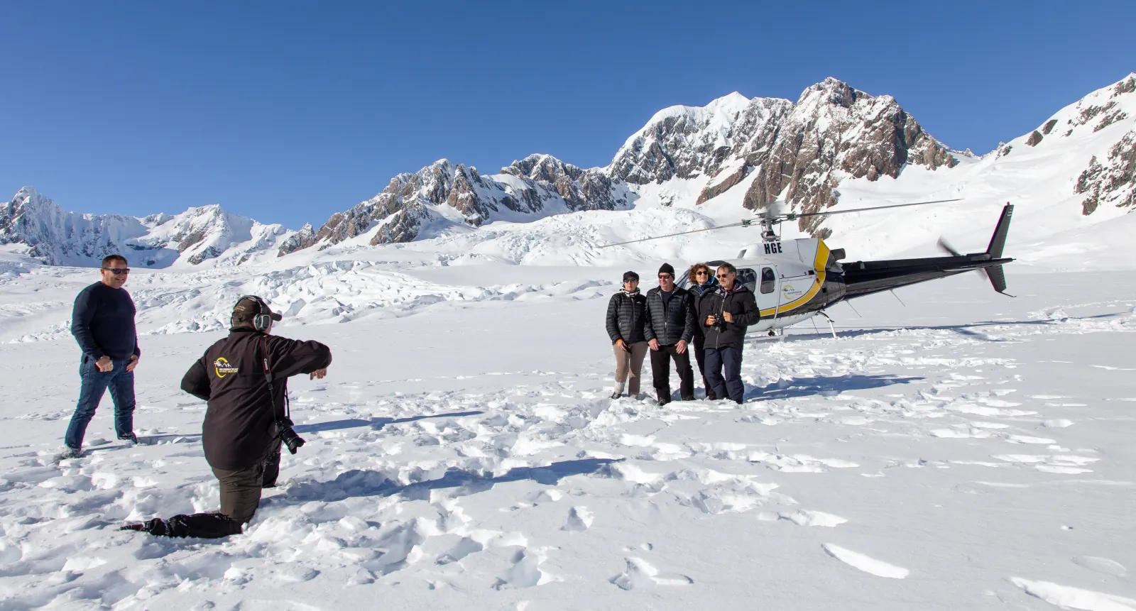 Visitors and a photographer on a wide snowfield near a parked yellow and black helicopter