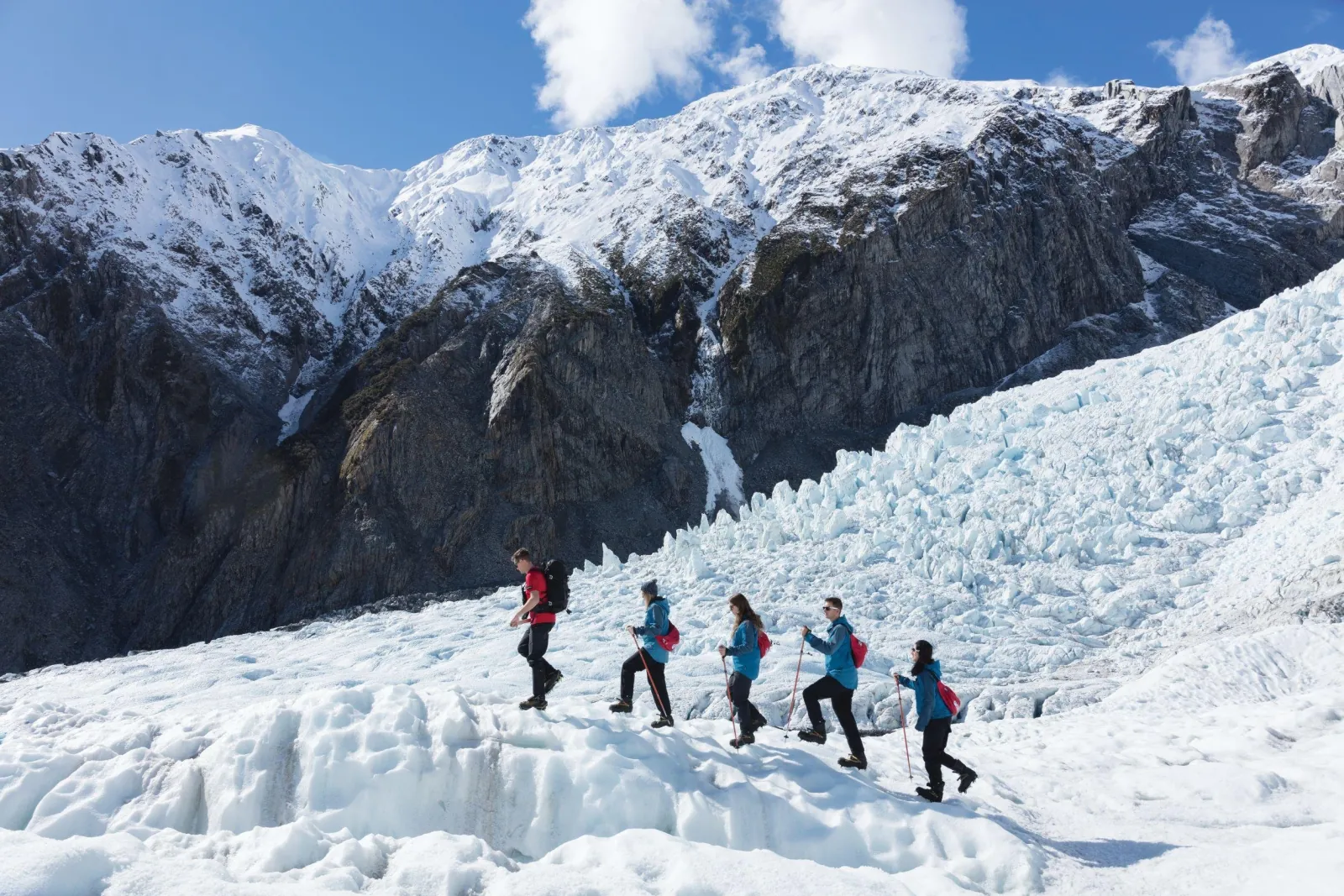 Five hikers walking in a line across rugged glacier ice beneath a snowy mountain