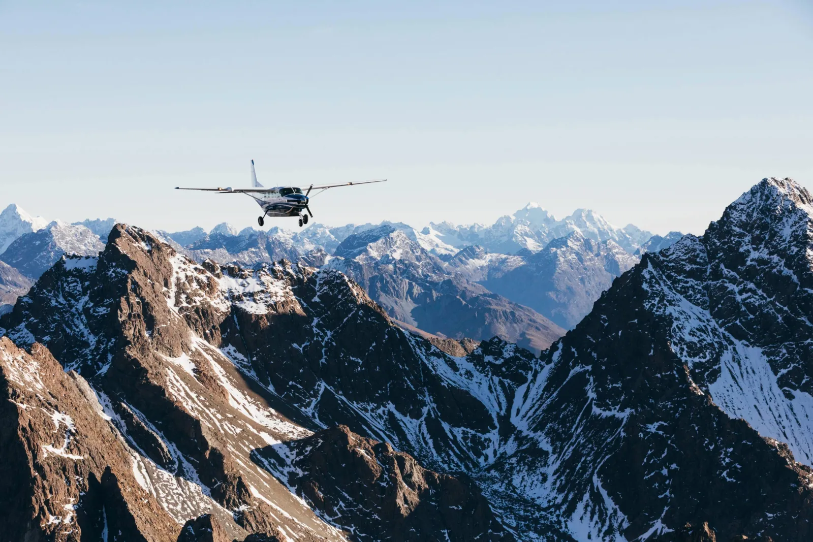 Small fixed-wing aircraft flying over rocky, snow-dusted ridges with higher peaks beyond