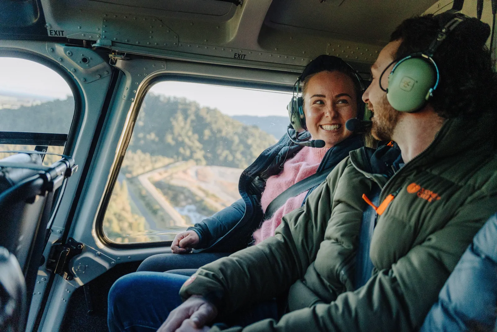 Two passengers in headsets smiling in a helicopter cabin with forest visible outside
