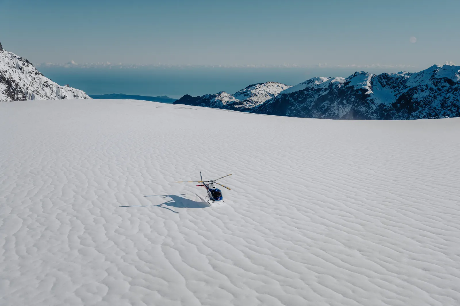 Lone helicopter on a rippled snow plateau with the sea and ranges beyond