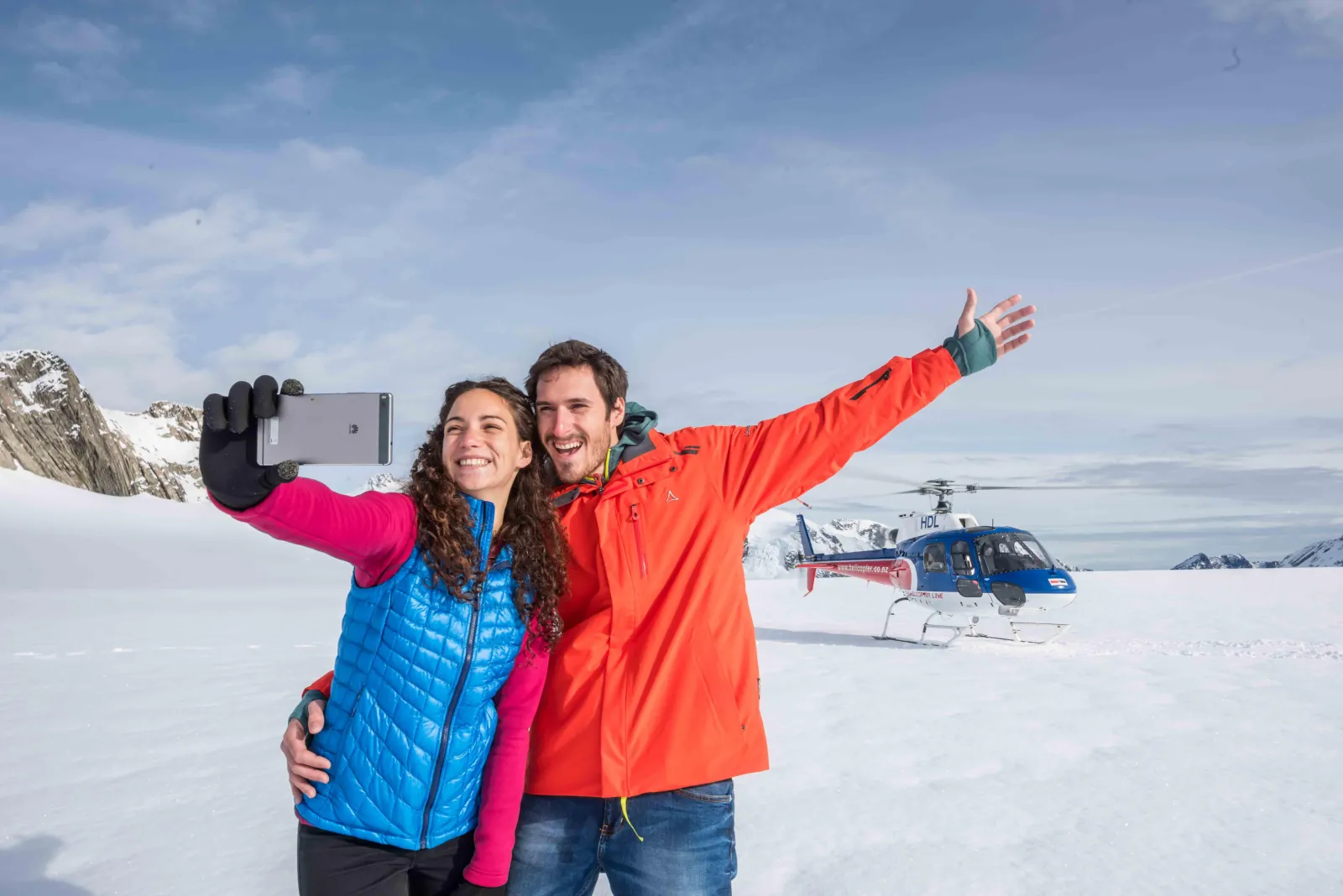 Couple taking a selfie on snow with a blue helicopter parked behind them